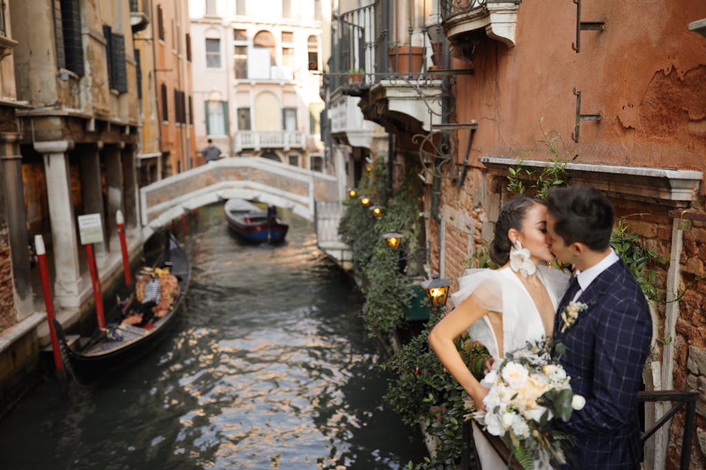 Bride and groom share a kiss beside a picturesque canal in Venice, Italy, with a gondola in the background.