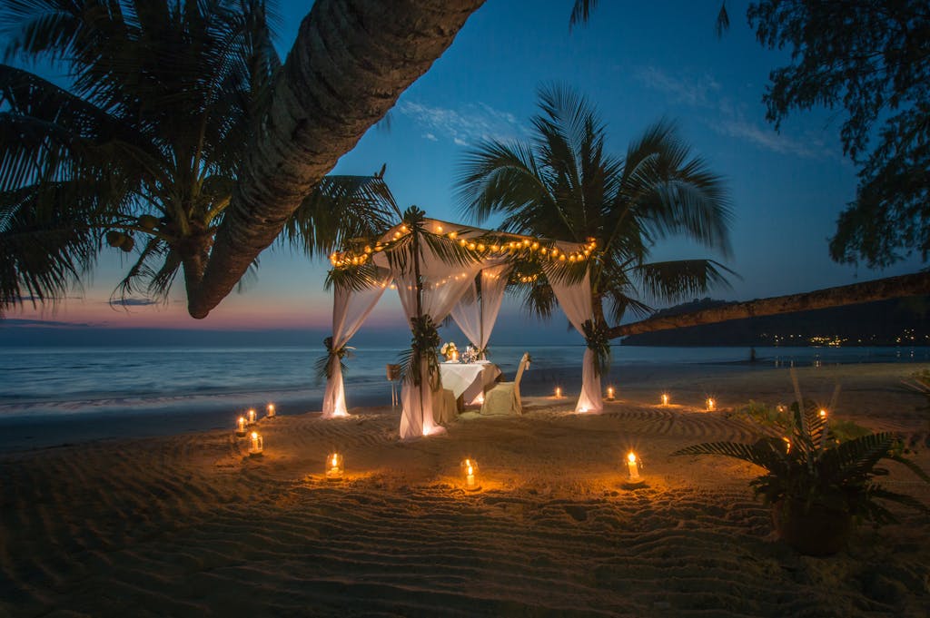 A picturesque romantic dinner setup under a canopy on a Thai beach at twilight, surrounded by candles.